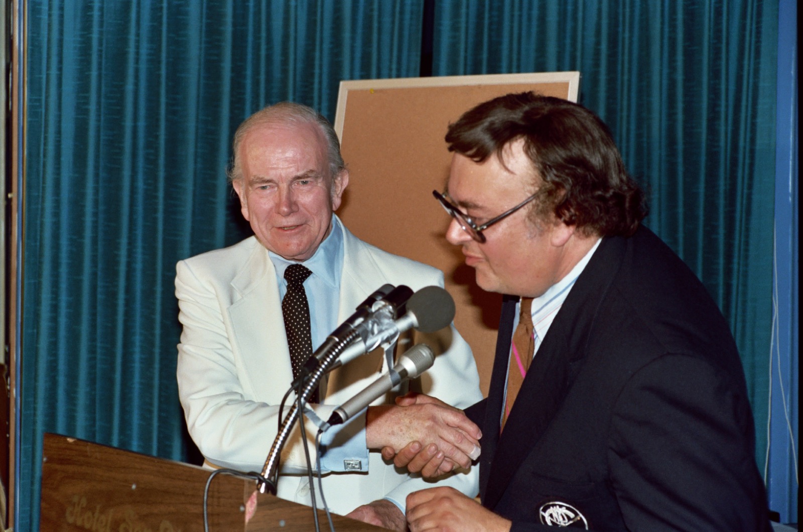 Milton Caniff in a white suit shaking hands at a convention podium