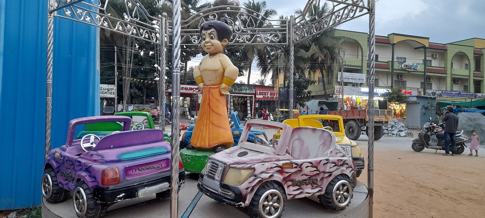 Chhota Bheem statue standing on a kiddie carousel ride surrounded by toy cars on an Indian street