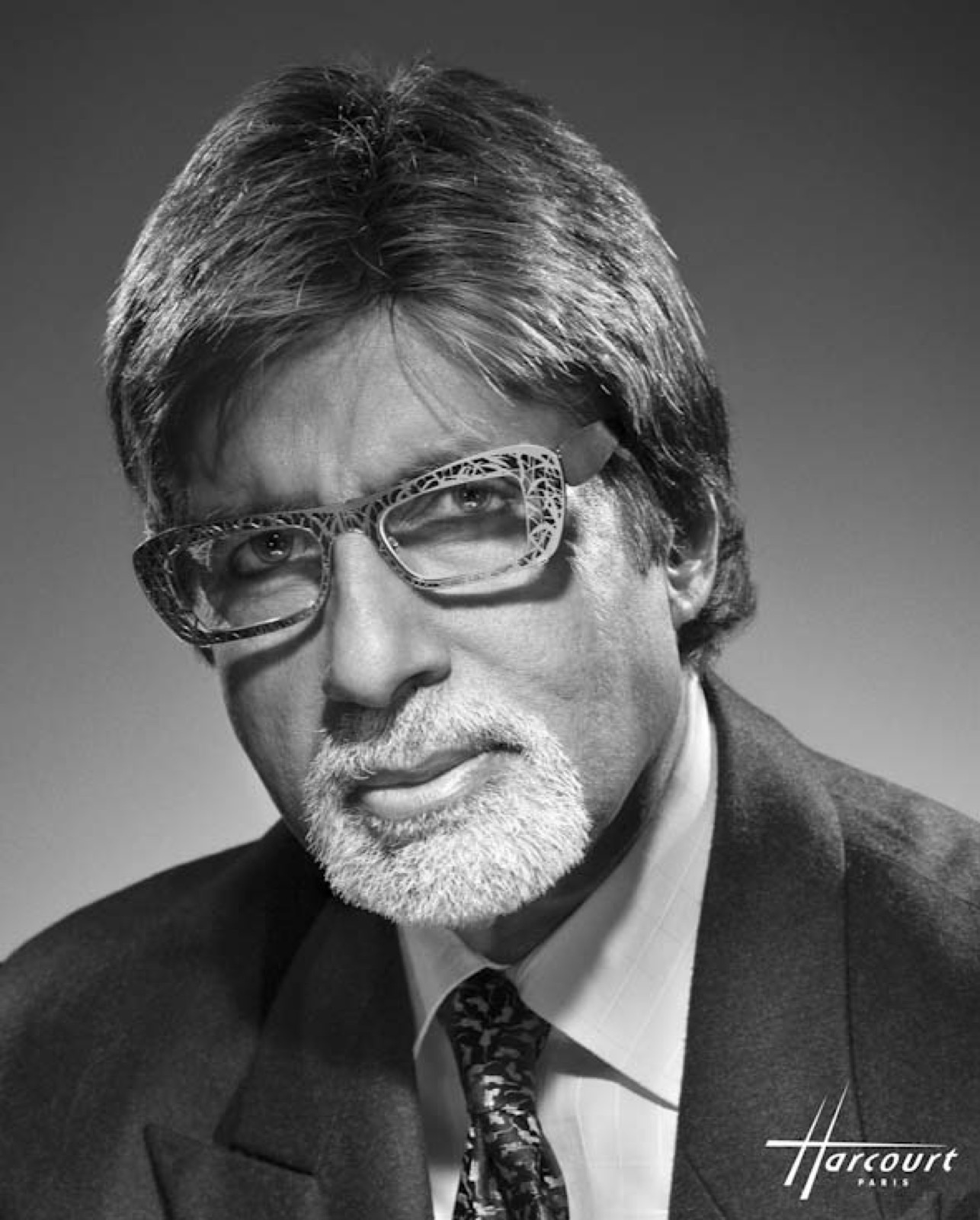 Black and white studio portrait of Amitabh Bachchan wearing glasses and a dark suit
