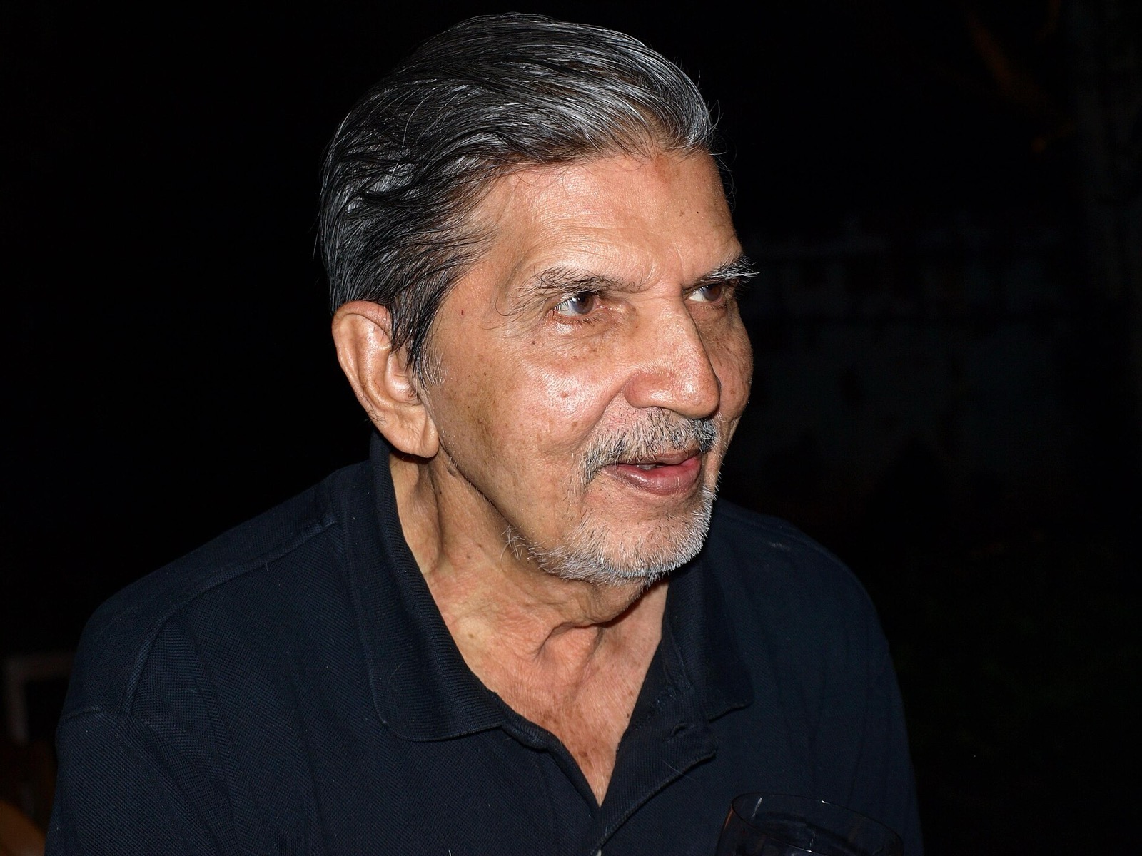 Elderly man with swept-back grey hair in a dark polo shirt, smiling at night