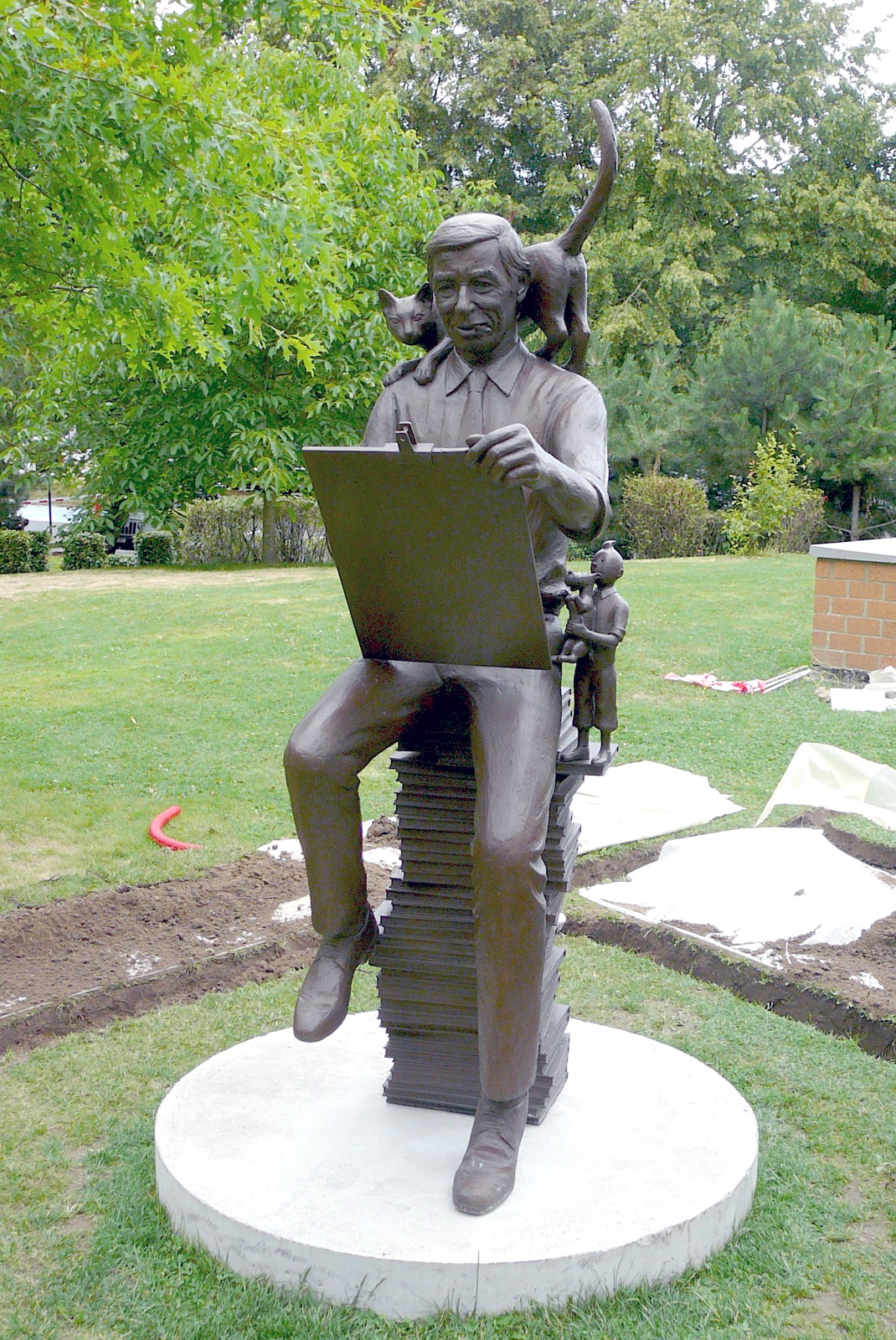 Bronze statue of Herge seated on stacked albums, drawing, with a cat on his shoulders and a small Tintin figure at his side