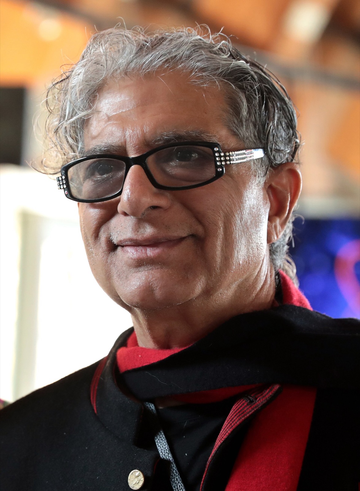 Deepak Chopra smiling, wearing black-rimmed glasses and a red scarf