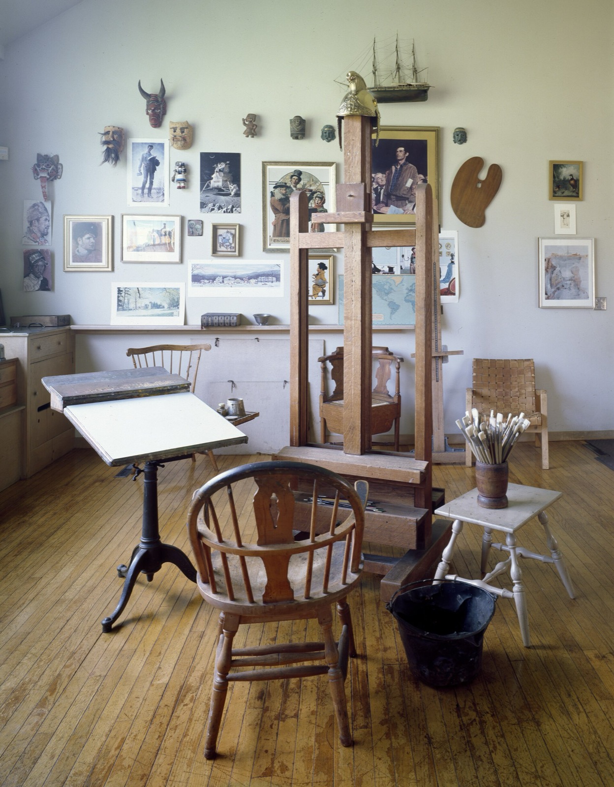 Studio interior with a tall wooden easel, drafting table, brushes and framed prints on the wall