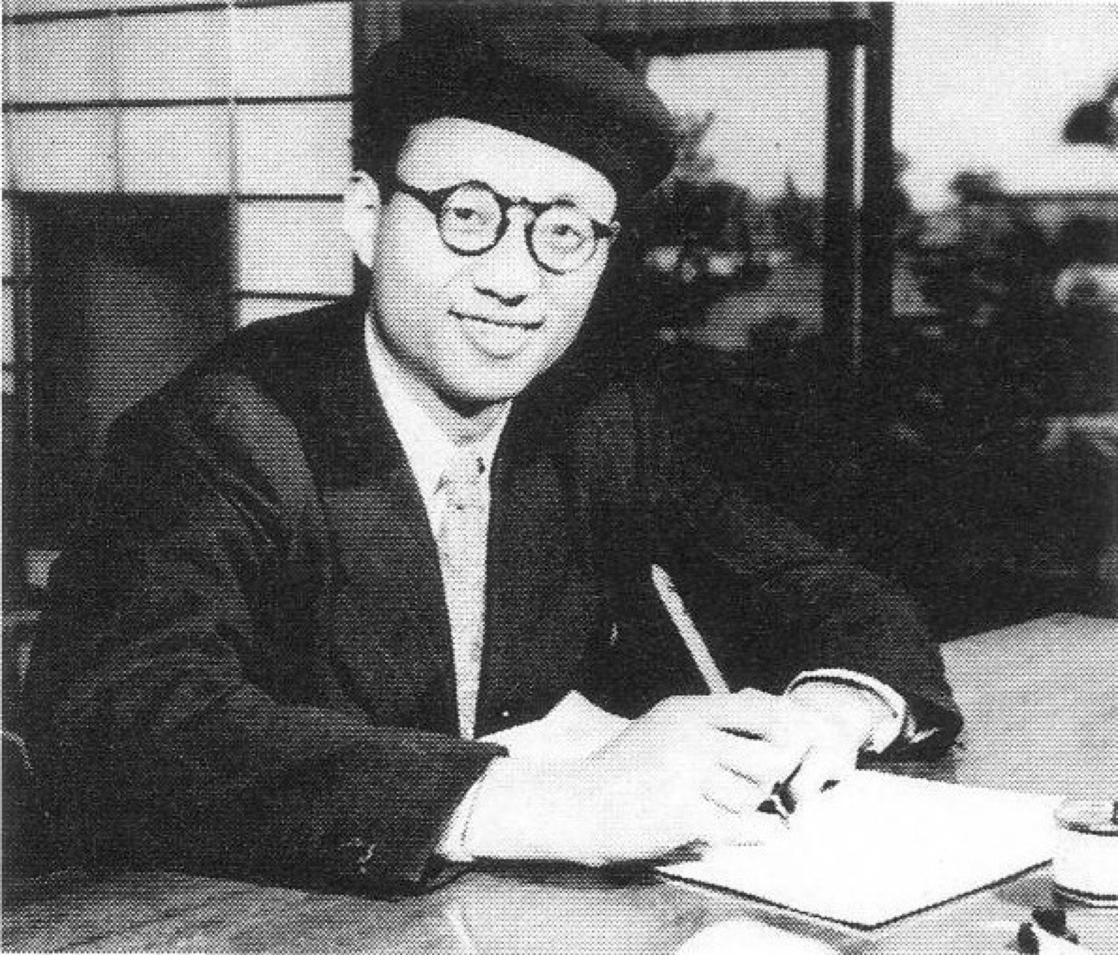 Black and white photo of a young Osamu Tezuka in a beret and round glasses, pen in hand at a desk