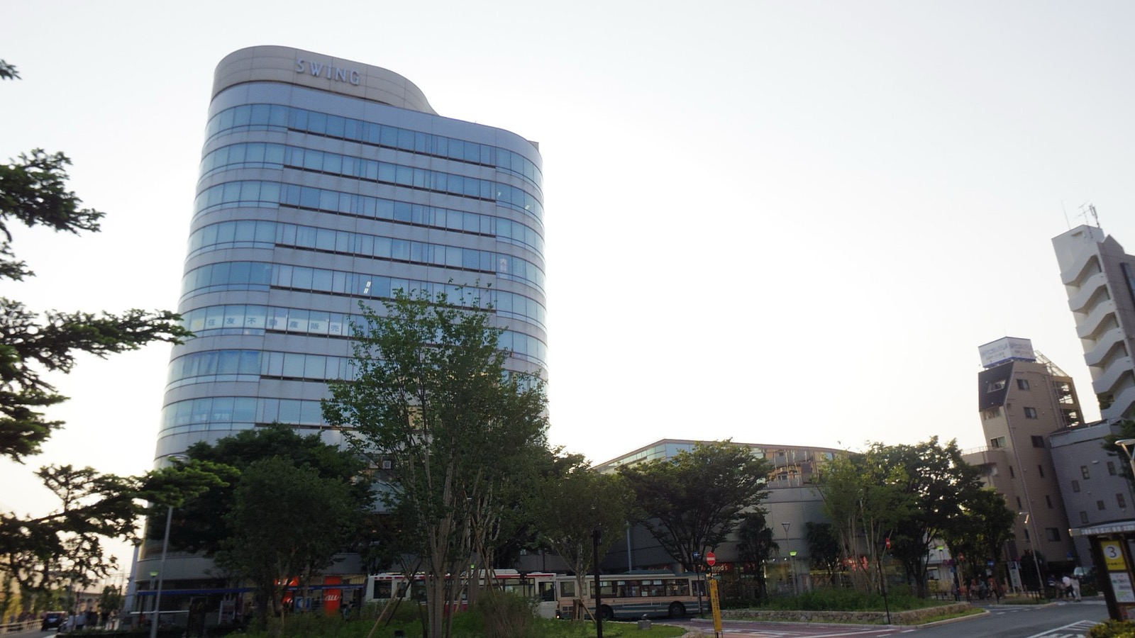 Curved glass office tower with a SWING sign rising above trees and buses